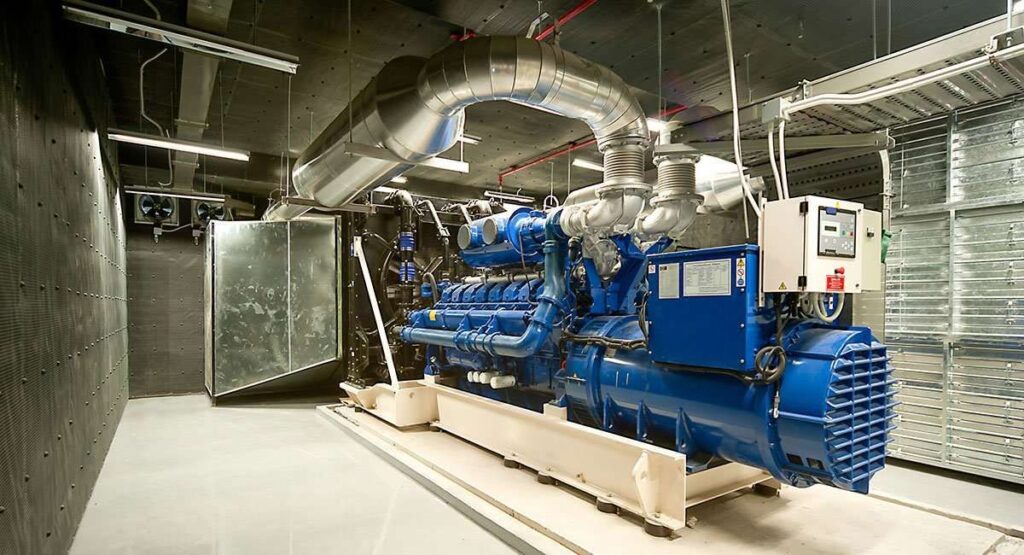 One of blue diesel generators housed in an industrial setting, with ventilation ducts and piping overhead.